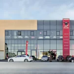 Otoclic car dealership with a glass storefront, beige upper panel, and a tall red sign on the right.