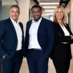 Three business professionals pose confidently in a modern office hallway, two men in navy suits and a woman in a black blazer smiling at the camera.