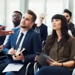 experts-de-coaching-en-entreprise-2026-1200x502 1 - Agence 47 Group of professionals in a conference room listening to a presenter, some taking notes on tablets and notebooks.