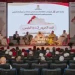 Panel discussion on a stage in a large auditorium; five men in traditional attire seated before a large screen with Arabic text and a map backdrop.