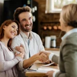 Smiling couple shakes hands with a person in a checkered jacket across a wooden counter, holding papers on the desk