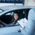 Woman driver smiling as a man presents a contract on a clipboard at her car window.