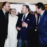 Four men in formal attire converse at an indoor event; one wears a white traditional robe, the others in dark suits, with flowers in the background.