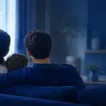 Back view of a family of three cuddled on a blue sofa watching television in a dim living room with blue lighting.