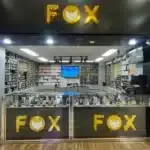 Electronics store interior with a black wall and a large yellow FOX logo above a glass display case filled with smartphones.