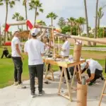 Group of people in white shirts assemble a long tubular frame outdoors among palm trees.