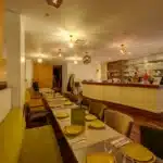 Restaurant dining room with a long table set for service, yellow plates and glasses, warm pendant lights overhead, and a bar area in the back.