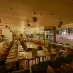 Warm-toned restaurant dining room with long rows of tables set for service and geometric pendant lights overhead.