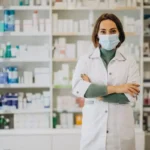 Pharmacist wearing a mask and white coat standing with arms crossed in a well-stocked pharmacy shelves behind her