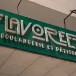 Green storefront sign with metallic letters reading 'FLAVOREZA Boulangerie et Patisserie' on a teal background behind the ceiling tiles .