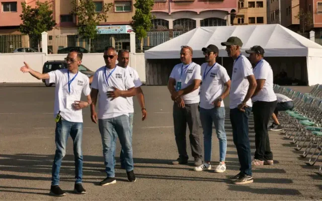 Group of event staff in white shirts standing in a parking lot, one man gesturing as he speaks near a white tent.