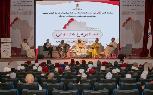 Panel discussion on a stage in a large auditorium; five men in traditional attire seated before a large screen with Arabic text and a map backdrop.