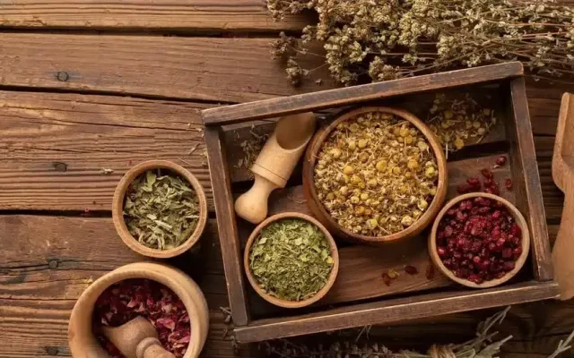 Top-down view of a wooden spice tray with bowls of dried herbs and spices on a rustic wooden table.