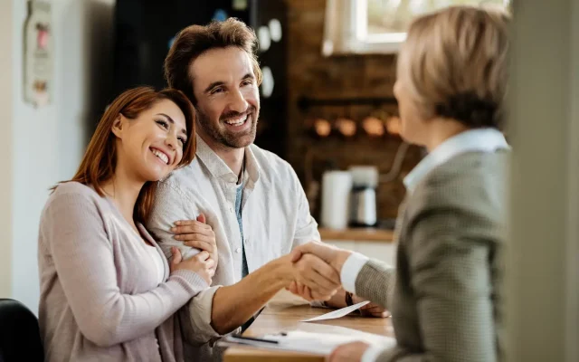 Smiling couple shakes hands with a person in a checkered jacket across a wooden counter, holding papers on the desk