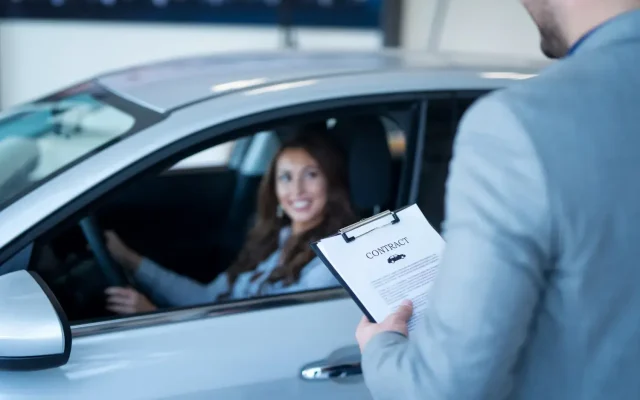 Woman driver smiling as a man presents a contract on a clipboard at her car window.
