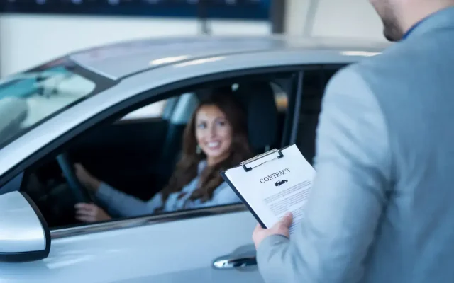Woman driver smiling as a man presents a contract on a clipboard at her car window.