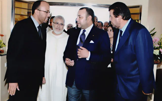 Group of men in suits chatting indoors, with one man in a white traditional robe and floral decor in the background.