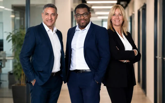 Three business professionals pose confidently in a modern office hallway, two men in navy suits and a woman in a black blazer smiling at the camera.