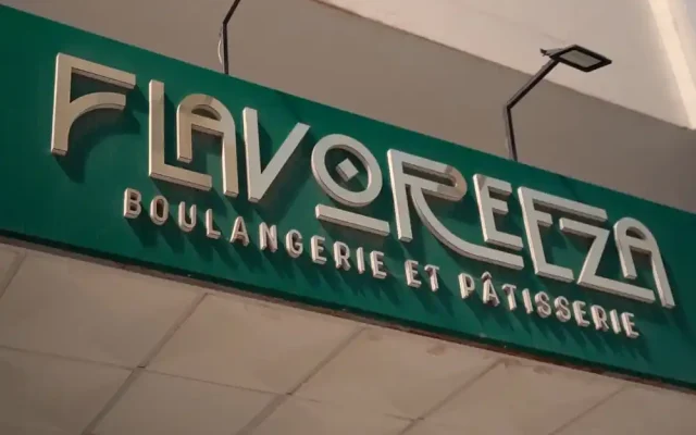Green storefront sign with metallic letters reading 'FLAVOREZA Boulangerie et Patisserie' on a teal background behind the ceiling tiles .