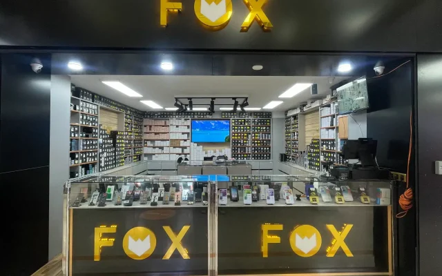 Electronics store interior with a black wall and a large yellow FOX logo above a glass display case filled with smartphones.