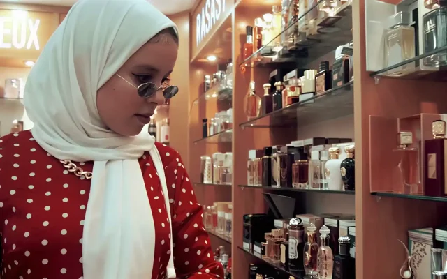 Woman in a white hijab and red polka-dot dress browsing perfume bottles on glass shelves in a store door-side shop lighting.