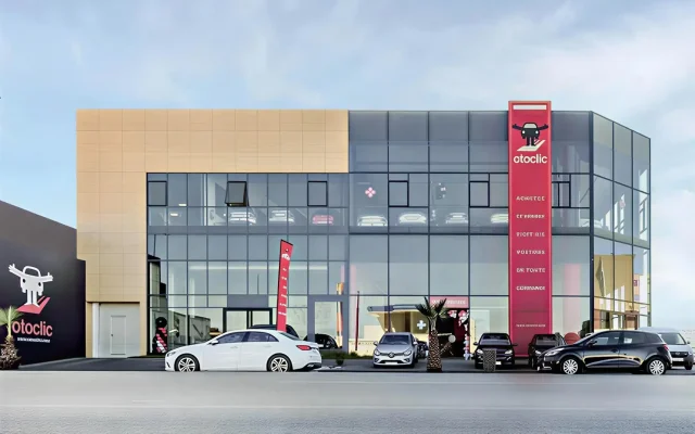 Otoclic car dealership with a glass storefront, beige upper panel, and a tall red sign on the right.