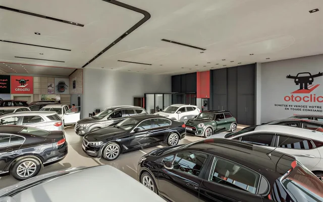 Indoor car showroom with multiple cars on display and Otoclic branding on the wall, bright showroom lighting above.