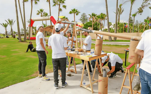 Group of people in white shirts assemble a long tubular frame outdoors among palm trees.