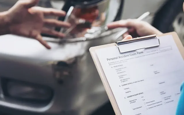 Clipboard with a Personal Accident Claim Form held near a damaged car involved in a collision. The scene shows hands reaching toward the vehicle for discussion or documentation.]?