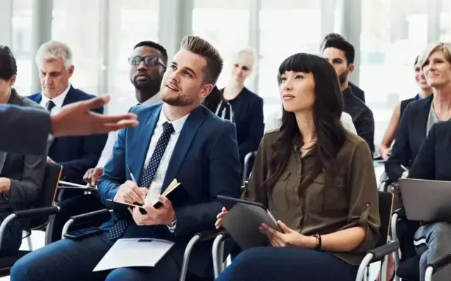 Group of professionals in a conference room listening to a presenter, some taking notes on tablets and notebooks.