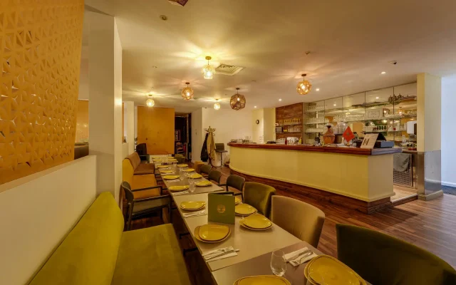 Restaurant dining room with a long table set for service, yellow plates and glasses, warm pendant lights overhead, and a bar area in the back.