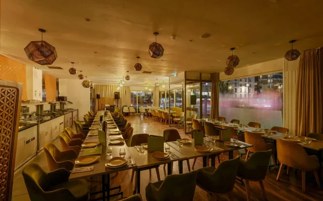 Warm-toned restaurant dining room with long rows of tables set for service and geometric pendant lights overhead.