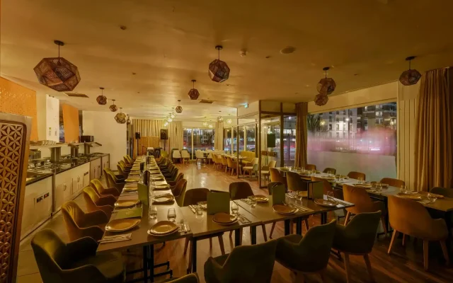 Warm-toned restaurant dining room with long rows of tables set for service and geometric pendant lights overhead.