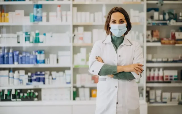 Pharmacist wearing a mask and white coat standing with arms crossed in a well-stocked pharmacy shelves behind her
